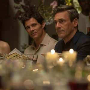 Four people—your friends & neighbors—sit closely together at a candlelit dinner table, holding menus and looking attentively forward. The table is elegantly set, and soft lighting creates a warm, intimate atmosphere.