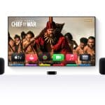 A TV displays the Apple TV home screen with the show "Chief of War" featuring Jason Momoa. Two black speakers stand on either side, set for perfect Apple TV audio sync, all arranged on a white surface. The Apple logo is in the corner.