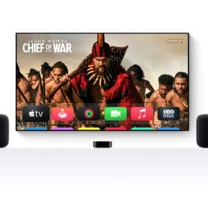 A TV displays the Apple TV home screen with the show "Chief of War" featuring Jason Momoa. Two black speakers stand on either side, set for perfect Apple TV audio sync, all arranged on a white surface. The Apple logo is in the corner.