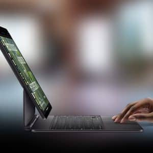 A hand types on an iPad external keyboard attached to a tablet, which is propped up on a desk. The background is blurred, and an Apple logo is visible in the bottom right corner.
