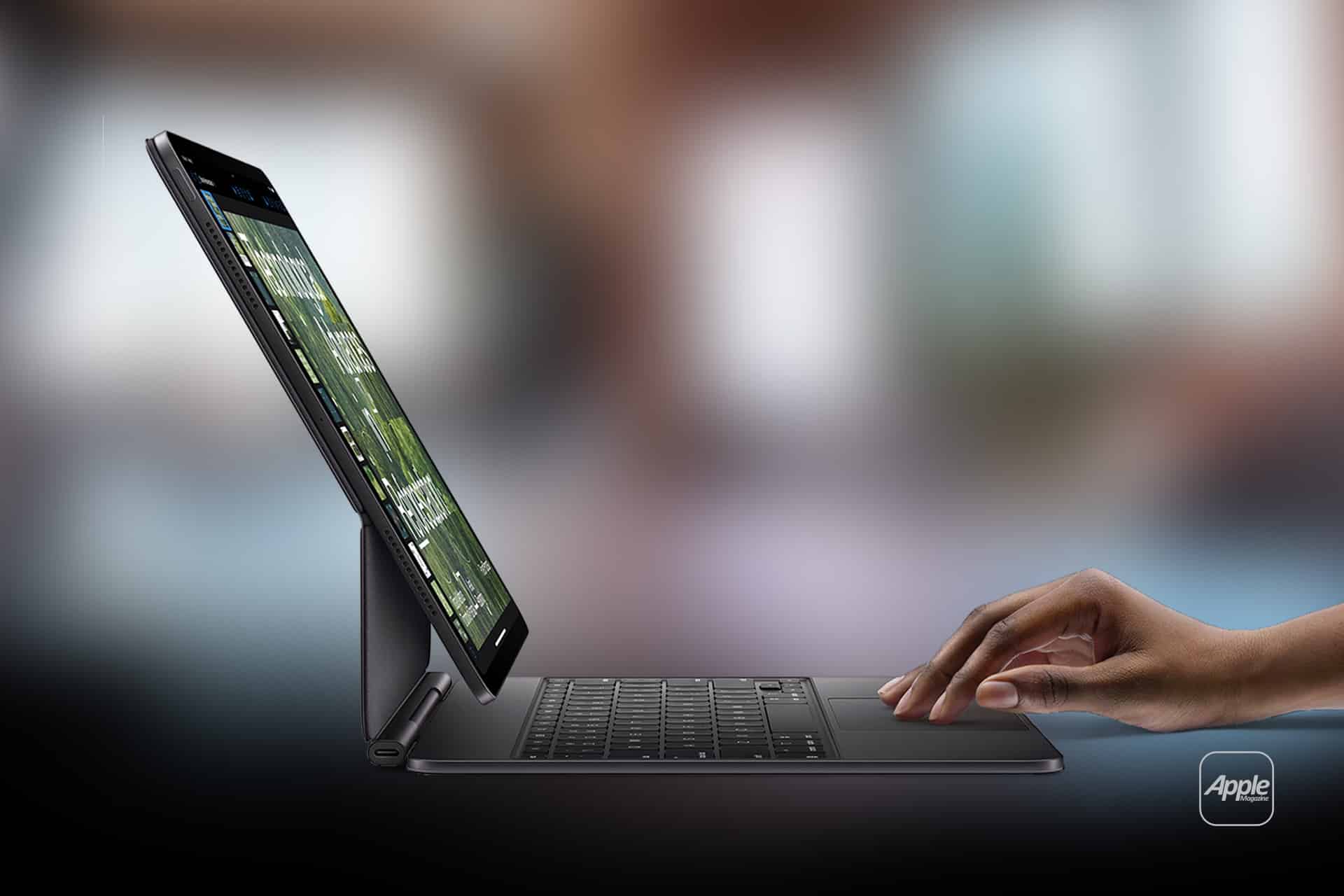 A hand types on an iPad external keyboard attached to a tablet, which is propped up on a desk. The background is blurred, and an Apple logo is visible in the bottom right corner.