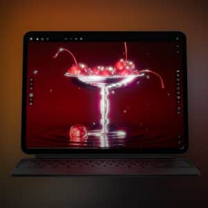 A tablet with a keyboard displays a digital image of cherries in a glass dessert dish against a deep red background. The Apple logo appears in the bottom right corner, and editing tools are visible on the iPad XDR monitor screen.