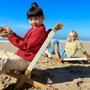 Two children sit on lounge chairs at the beach. The child in front, in a red sweater, smiles and holds sand—curious like young minds exploring machine learning. Behind them, the ocean and blue sky stretch into the distance.