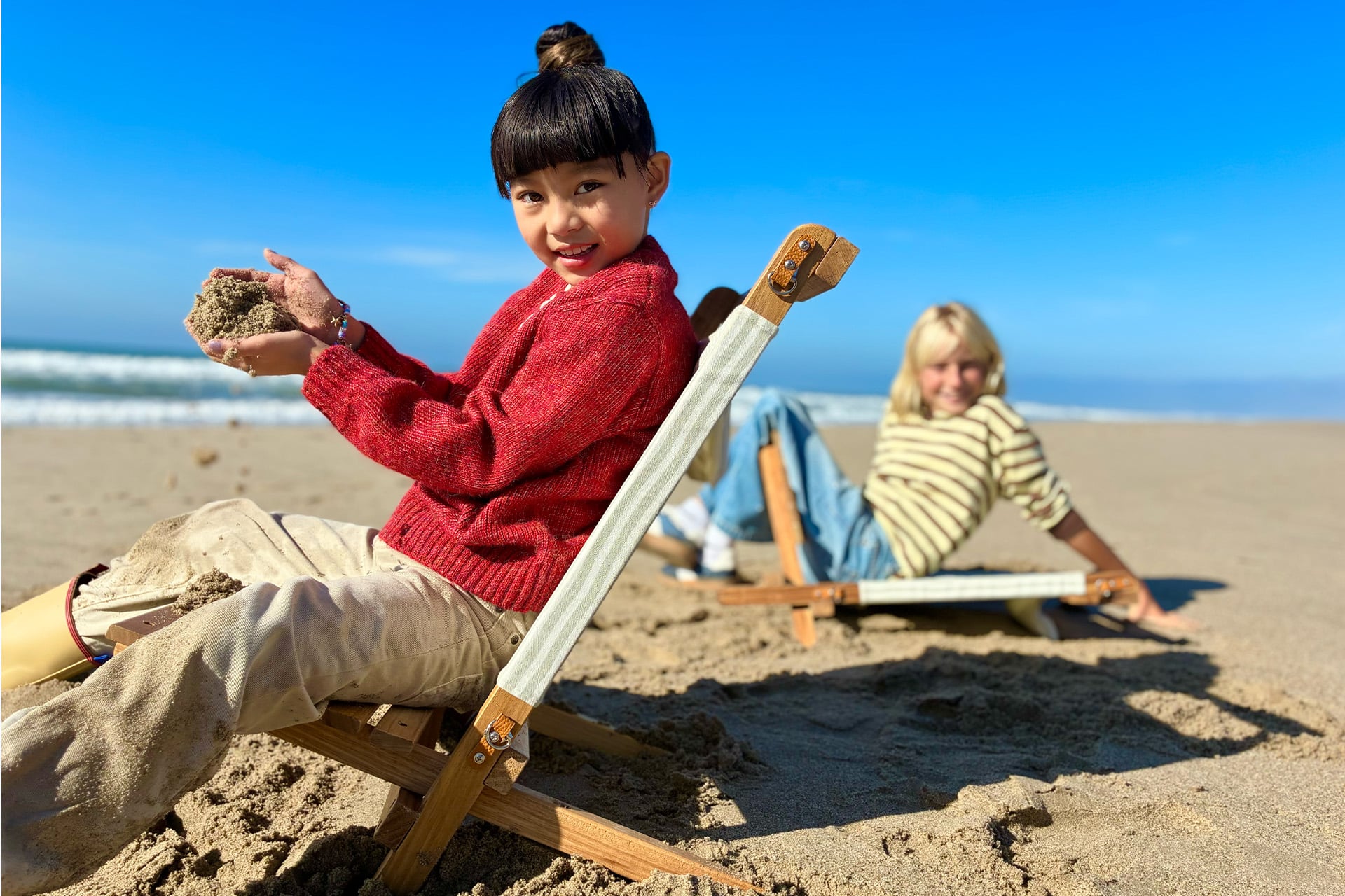 Two children sit on lounge chairs at the beach. The child in front, in a red sweater, smiles and holds sand—curious like young minds exploring machine learning. Behind them, the ocean and blue sky stretch into the distance.