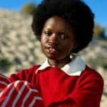 A person with an afro hairstyle, dressed in a red sweater with a white collar and red-and-white striped pants, sits on a sandy beach under blue skies and lush vegetation, pondering the possibilities of machine learning.