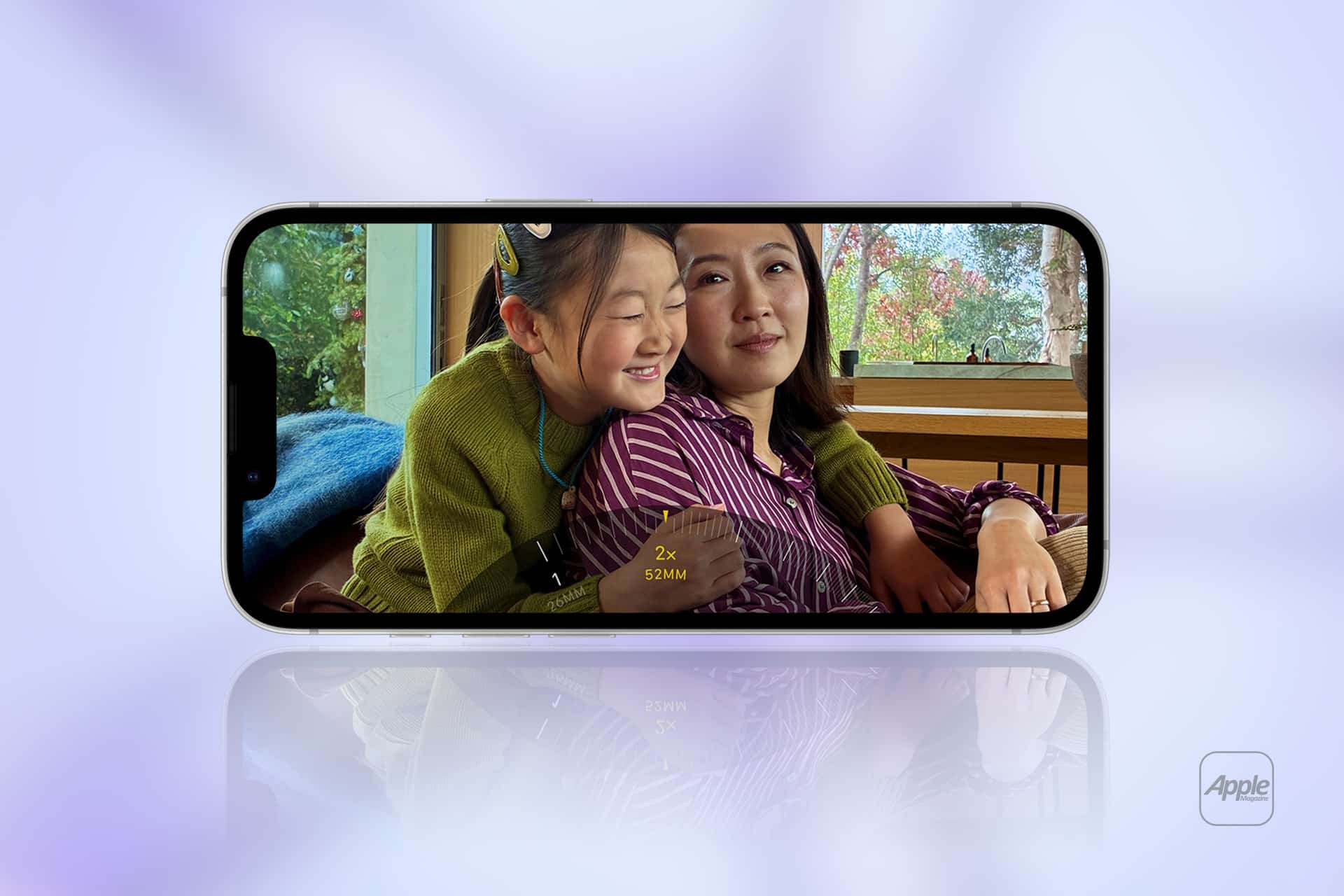 A smartphone displaying a photo of two people sitting together indoors, with one person smiling and hugging the other. The phone rests on a reflective surface with a blurred lavender background and an Apple logo in the corner.