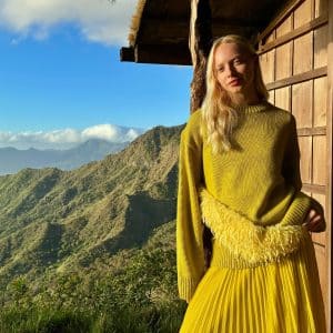 A woman in a yellow sweater and pleated skirt stands by a wooden building, with sunlit mountains behind her and a blue sky above—captured using Apple Core AI and machine learning for vivid detail and clarity.