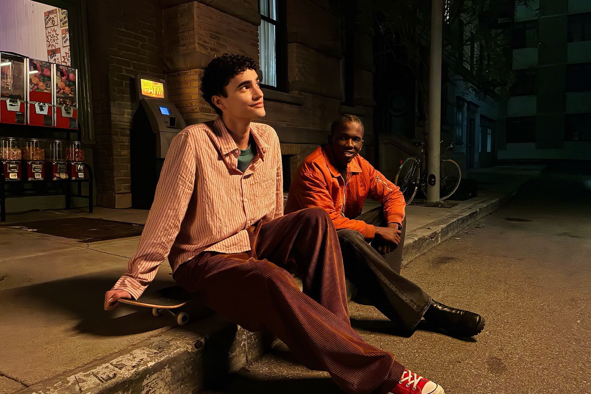Two young men sit on a curb at night in an urban setting. One leans back on a skateboard, smiling thoughtfully, while the other sits nearby. The street is dimly lit with vending machines and hints of machine learning in the city’s tech-driven ambiance.