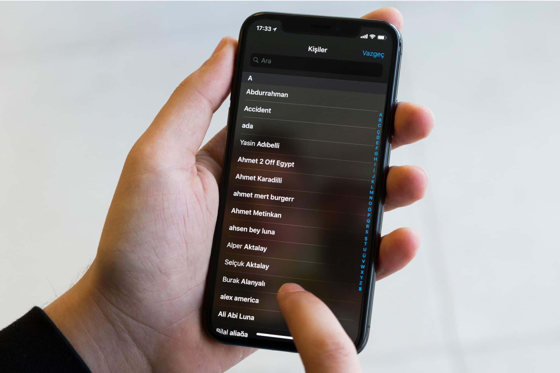 A person holds a smartphone displaying a contacts list in Turkish, their thumb scrolling through names—perhaps searching for duplicates to use the iPhone Contact Merge feature. The background is out of focus.