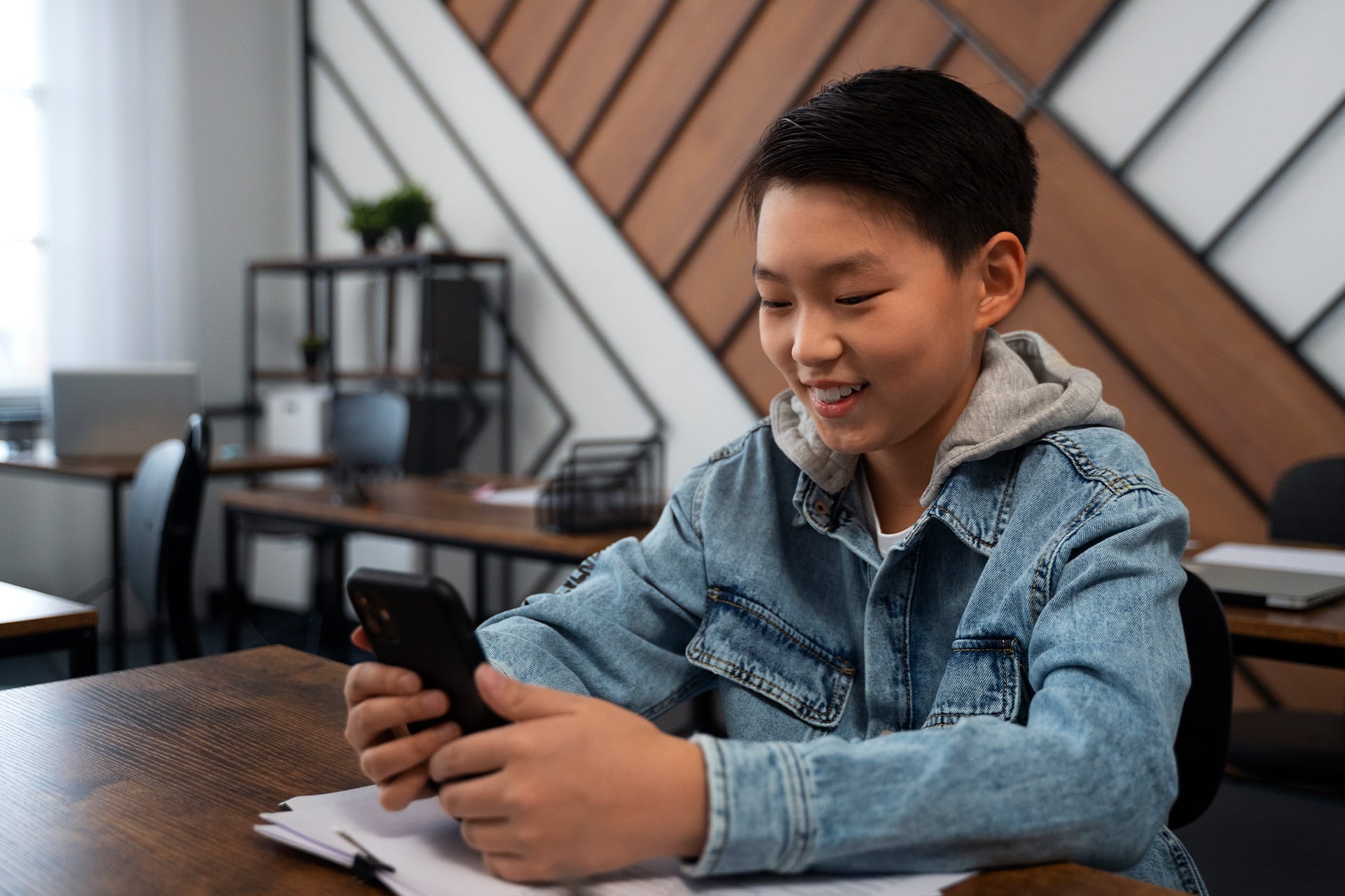 A smiling boy in a denim jacket sits at a desk in a modern classroom, engaging in school smartphone use while looking at his phone, with papers and books spread out before him.