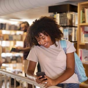 A young woman with curly hair and a backpack smiles while looking at her phone in a library, illustrating school smartphone use. Bookshelves and other people browsing books are visible in the background.