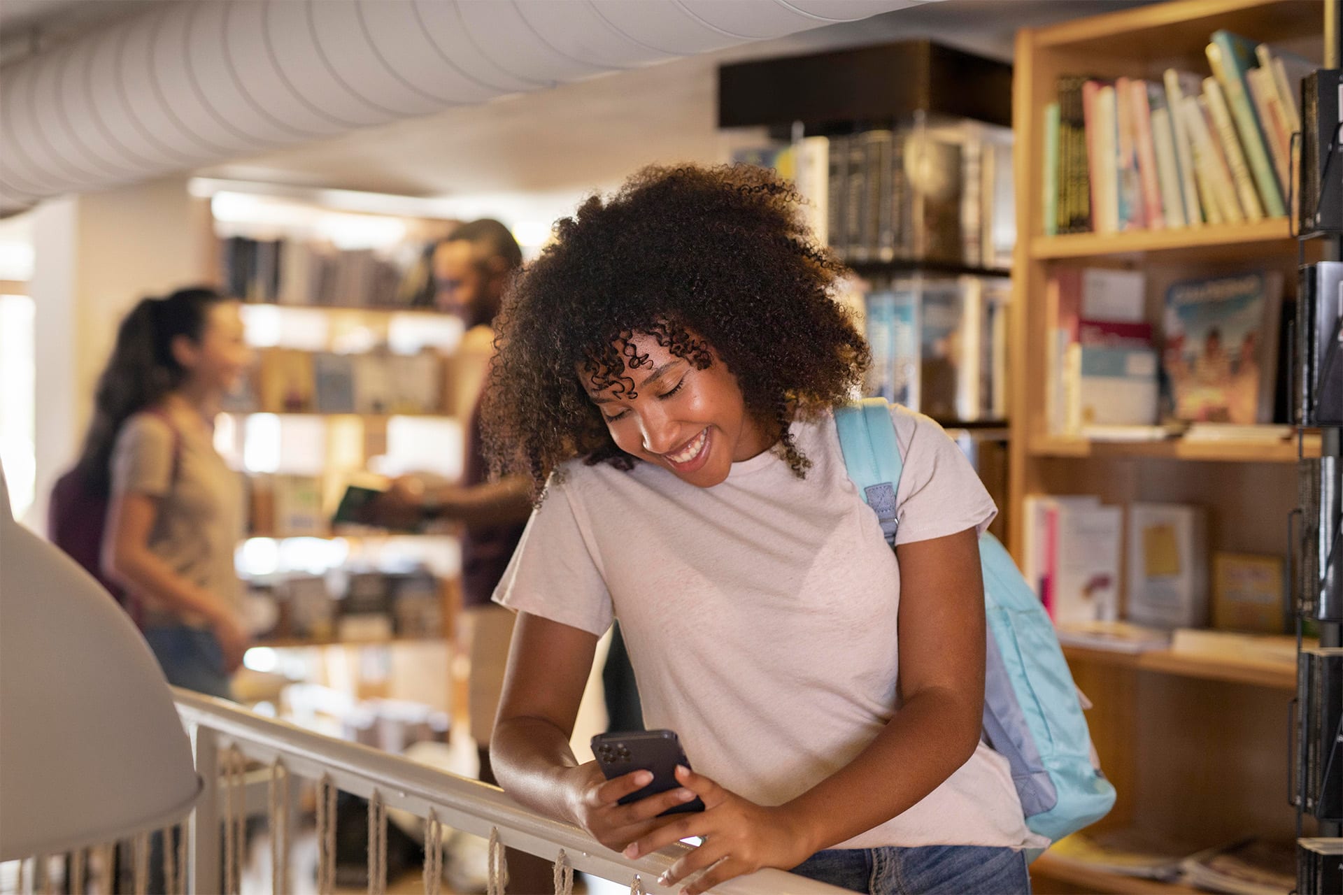 A young woman with curly hair and a backpack smiles while looking at her phone in a library, illustrating school smartphone use. Bookshelves and other people browsing books are visible in the background.