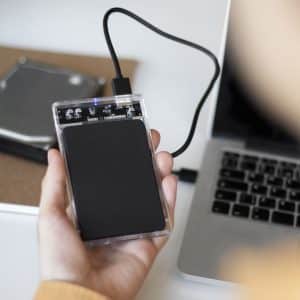 A person holds an external SSD setup connected by USB to a laptop. A notebook and an internal hard drive are on the desk in the background, highlighting a data transfer or backup process.