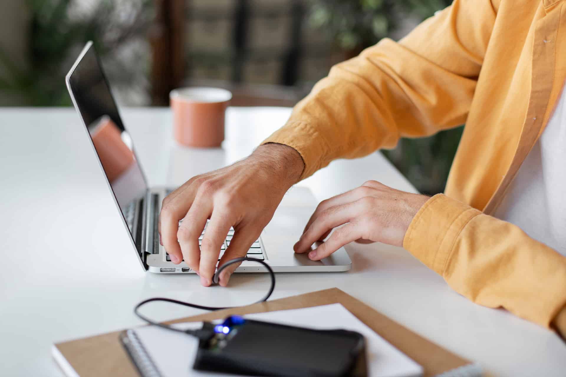 A person in a yellow shirt uses a laptop on a white desk, managing an external SSD setup. A notebook and a mug are also on the desk.