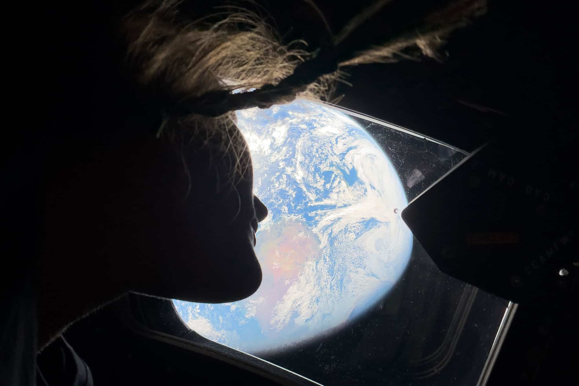 A person with floating hair looks out of a spacecraft window at Earth, which appears bright and detailed against the darkness of space—perfect inspiration for Artemis II wallpapers.