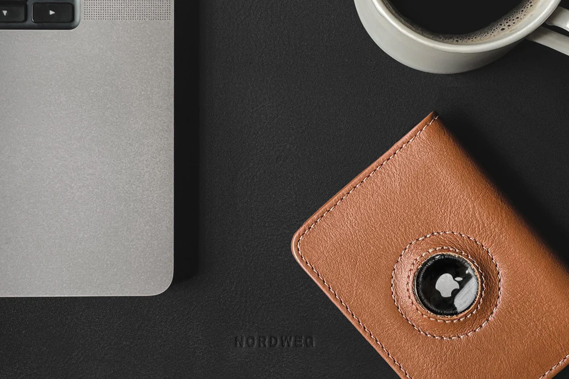 A close-up of a laptop, a brown leather wallet with an Apple AirTag, and a cup of black coffee on a dark desk highlights stylish AirTag accessory choices. The word “NORDWEQ” is faintly visible on the desk surface.