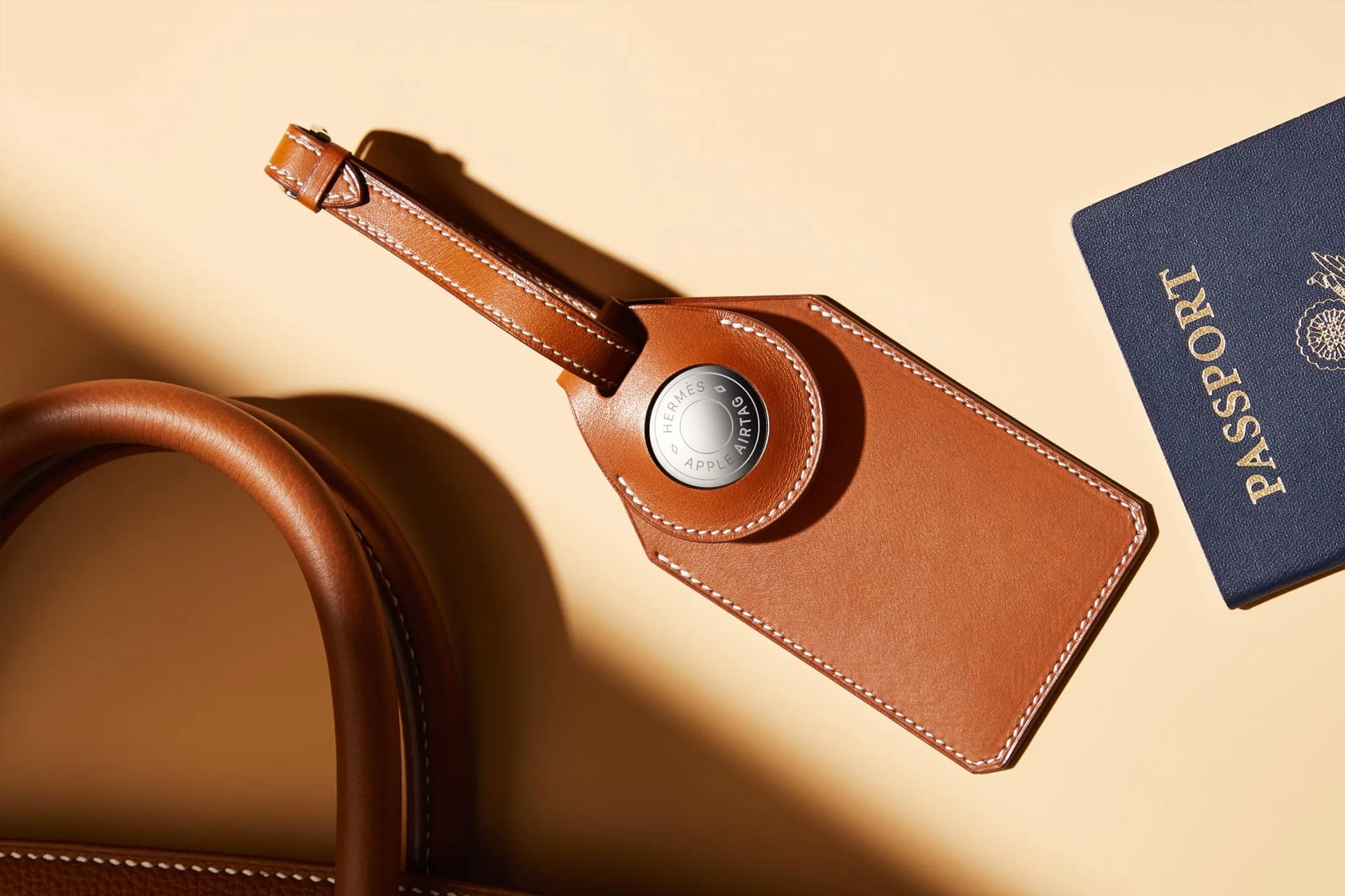 A brown leather luggage tag, one of many AirTag accessory choices, holds an Apple AirTag and rests beside a navy blue passport on a beige surface, with part of a matching leather bag visible.