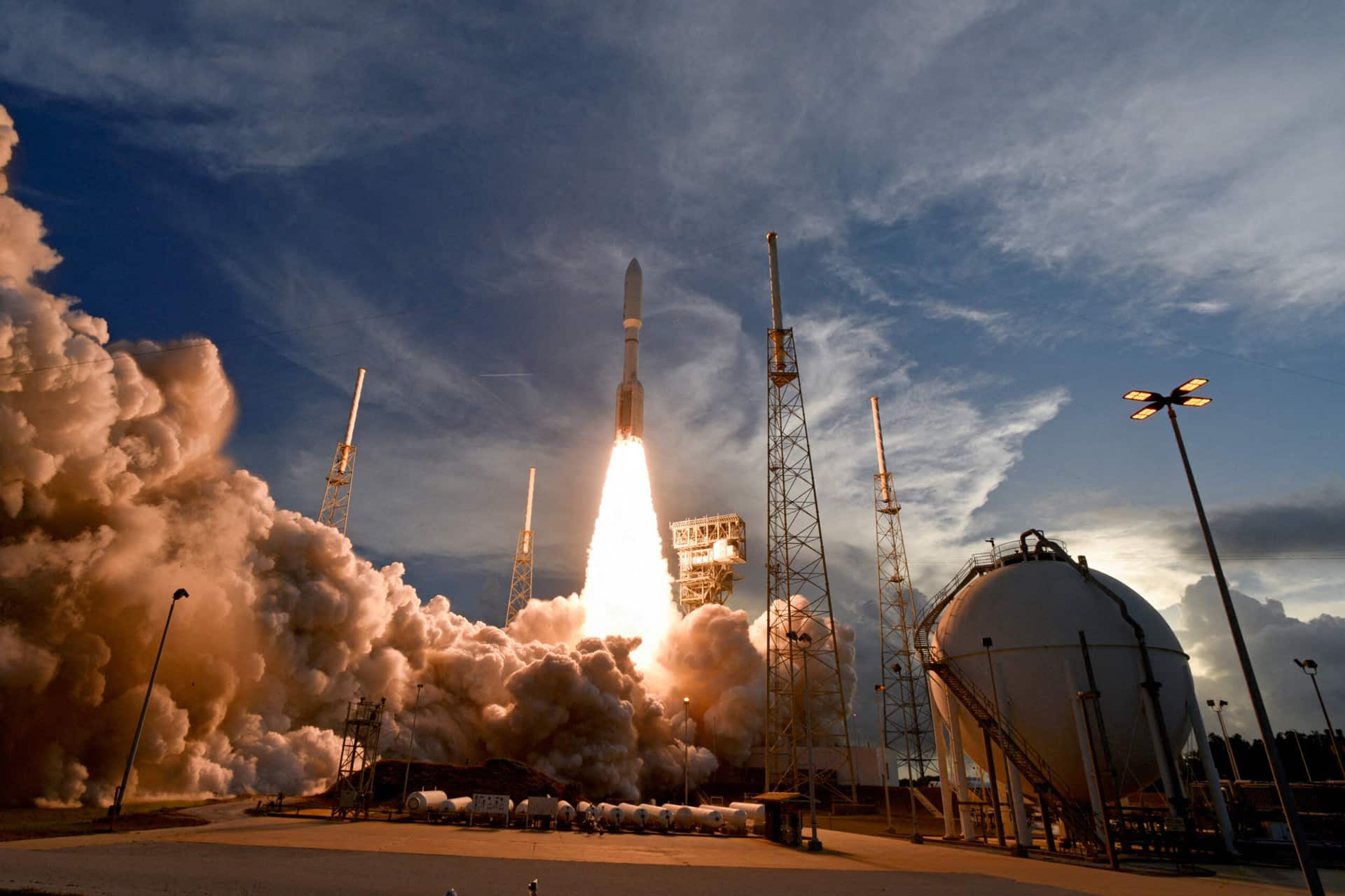 A rocket launches vertically from a launch pad, surrounded by clouds of smoke and flames, with tall support towers and a spherical storage tank under a partly cloudy sunset sky, preparing for its direct satellite mission.