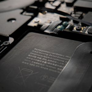 Close-up of the interior of an electronic device, showing a lithium-ion battery sourced from Apple battery suppliers, with safety warnings and components such as wiring, screws, and circuit boards. The text on the battery is partially visible.