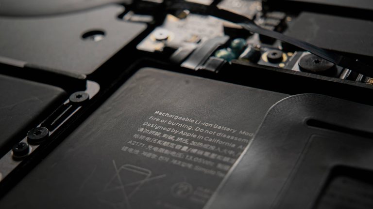 Close-up of the interior of an electronic device, showing a lithium-ion battery sourced from Apple battery suppliers, with safety warnings and components such as wiring, screws, and circuit boards. The text on the battery is partially visible.