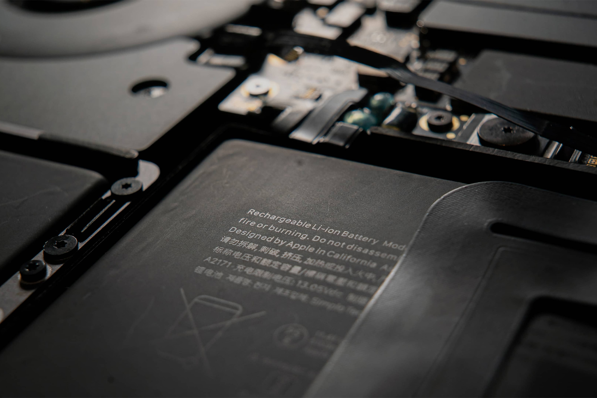 Close-up of the interior of an electronic device, showing a lithium-ion battery sourced from Apple battery suppliers, with safety warnings and components such as wiring, screws, and circuit boards. The text on the battery is partially visible.