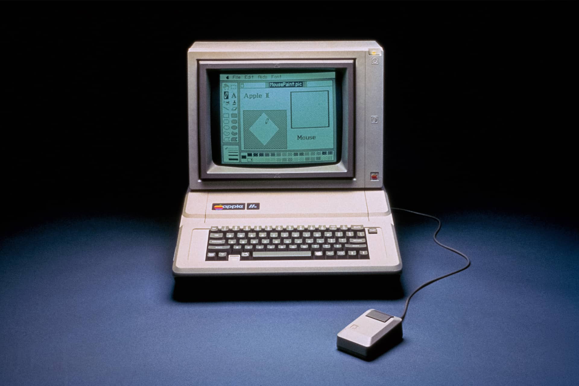 A vintage Apple II computer, famously co-created by Steve Wozniak, features a built-in monitor displaying an early graphical user interface with a window labeled "Apple I." A wired mouse rests to the right of the keyboard.
