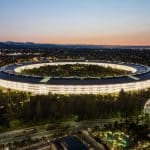 Aerial view of the large, circular Apple Park Visitor Center illuminated at dusk, surrounded by trees and roads, with a cityscape and mountains visible in the background.