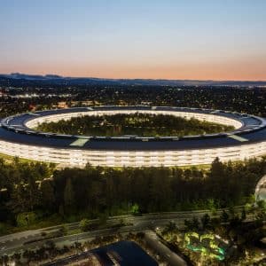 Aerial view of the large, circular Apple Park Visitor Center illuminated at dusk, surrounded by trees and roads, with a cityscape and mountains visible in the background.