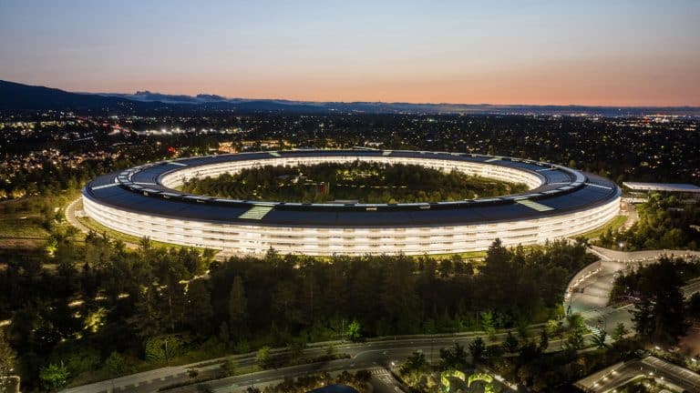 Aerial view of the large, circular Apple Park Visitor Center illuminated at dusk, surrounded by trees and roads, with a cityscape and mountains visible in the background.