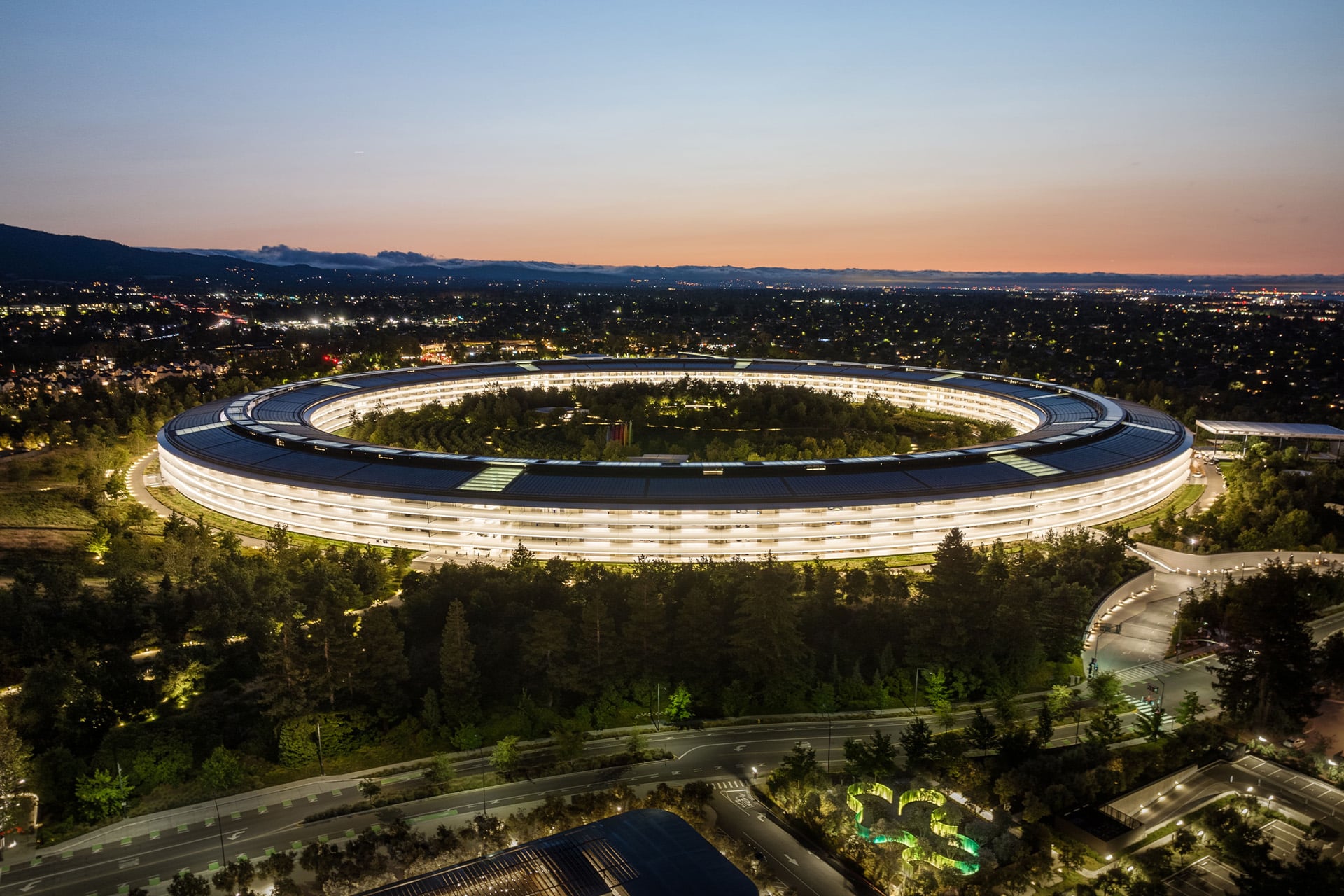 Aerial view of the large, circular Apple Park Visitor Center illuminated at dusk, surrounded by trees and roads, with a cityscape and mountains visible in the background.