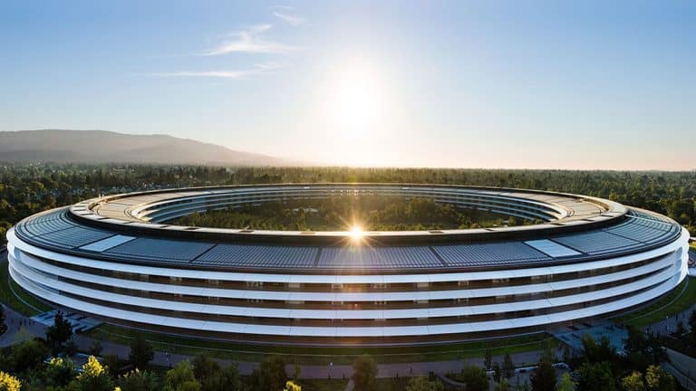 The image shows a large, circular modern building surrounded by trees, with the sun rising or setting in the background. The sleek, white architecture resembles Apple’s campus—an icon often seen in discussions about their Q2 earnings.