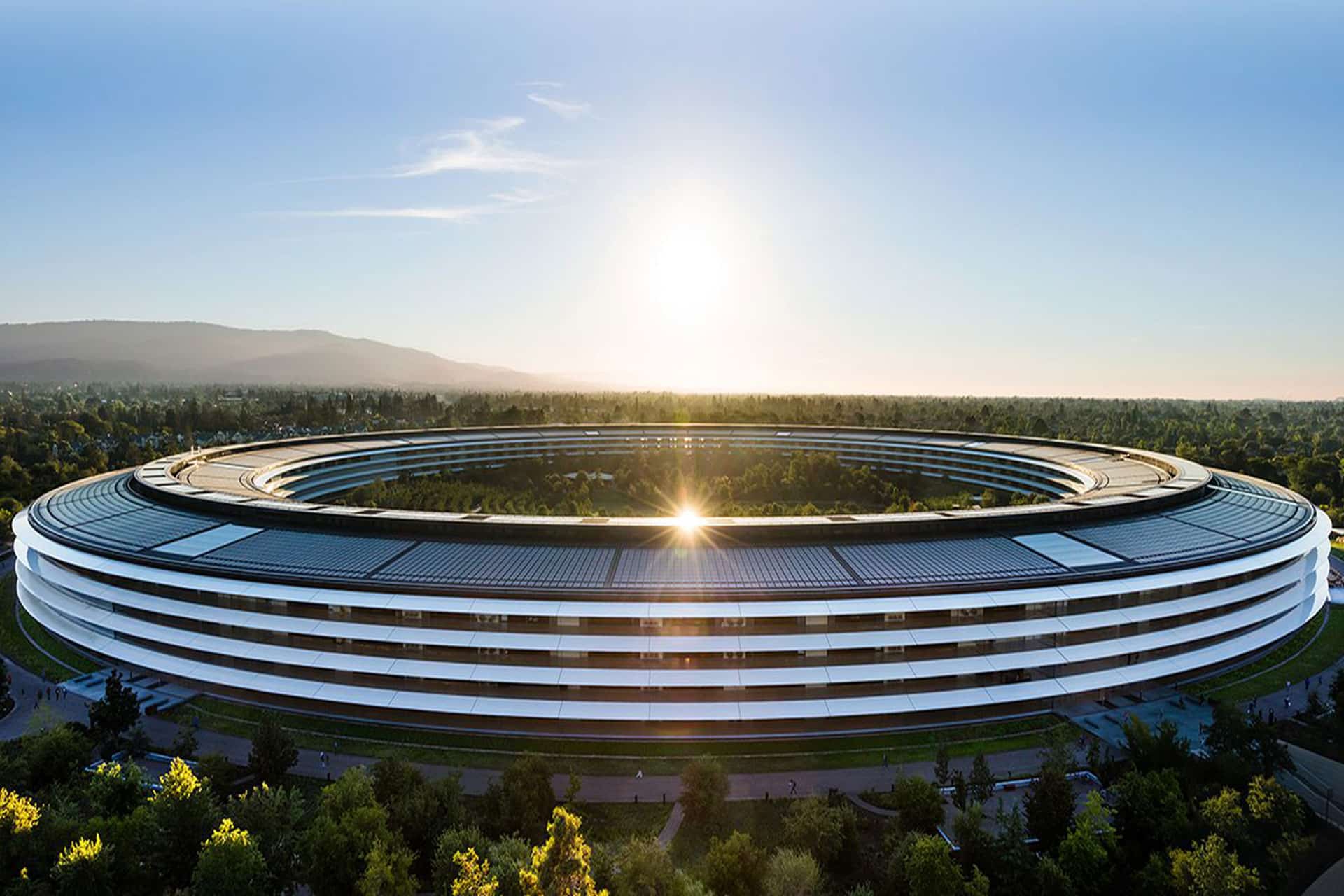 The image shows a large, circular modern building surrounded by trees, with the sun rising or setting in the background. The sleek, white architecture resembles Apple’s campus—an icon often seen in discussions about their Q2 earnings.