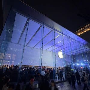 A large crowd gathers outside a brightly lit Apple Store with a glass facade at night, discussing Apple's cash reserve strategy beneath the illuminated logo prominently displayed above the entrance.