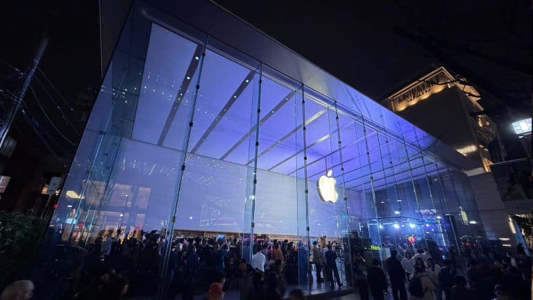 A large crowd gathers outside a brightly lit Apple Store with a glass facade at night, discussing Apple's cash reserve strategy beneath the illuminated logo prominently displayed above the entrance.