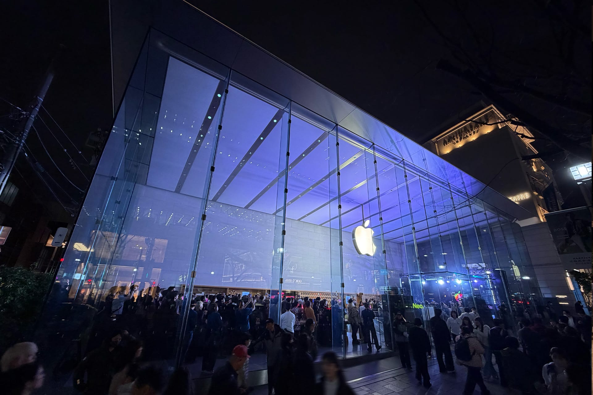 A large crowd gathers outside a brightly lit Apple Store with a glass facade at night, discussing Apple's cash reserve strategy beneath the illuminated logo prominently displayed above the entrance.
