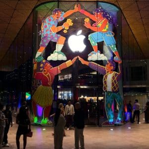 People stand outside an Apple Store at night, vibrant mural and glowing logo above them. The lively scene hints at more than tech—Apple’s success reflects a smart cash reserve strategy behind the colorful, bustling storefront. India Store.
