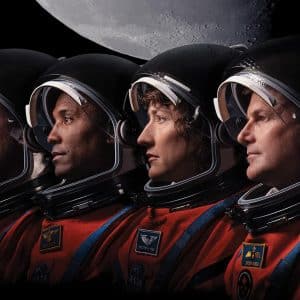 Four Artemis II astronauts wearing orange spacesuits and black helmets are shown in profile, looking to the right, with the gray surface of the Moon above them against a black background—ready for their mission as iPhones capture every moment.