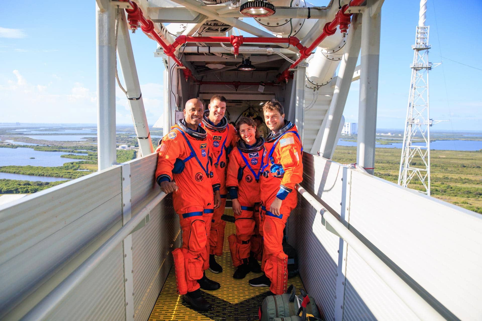 Four astronauts in bright orange space suits stand and smile together on a metal walkway high above the ground, with Artemis II iPhones capturing the moment amid green fields, water, and a launch pad visible in the background.