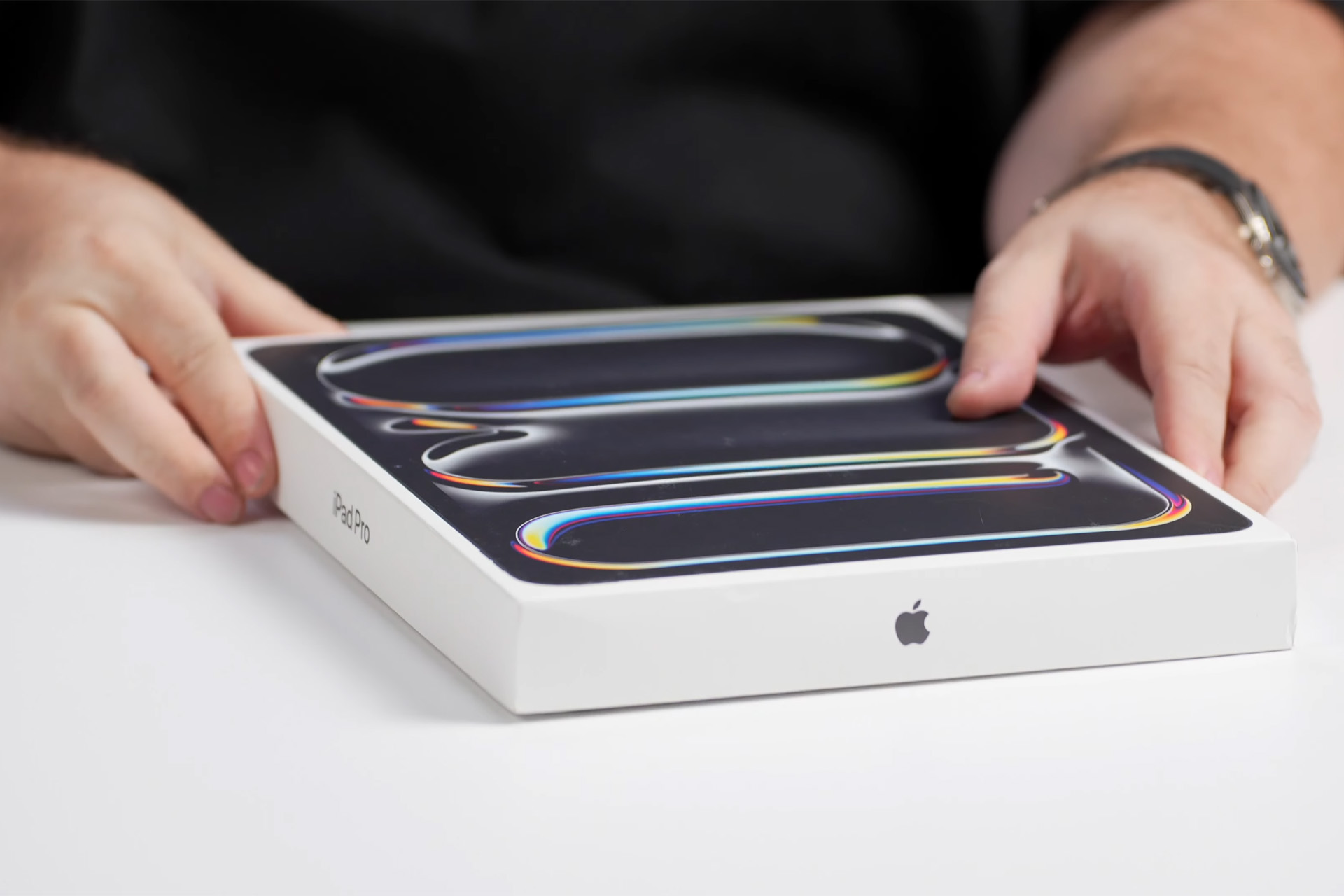 A person’s hands hold a sealed Apple iPad box on a white table, capturing the excitement of an Apple unboxing with colorful lines on the packaging and a visible Apple logo on the side.