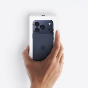 A hand holding a blue iPhone partially out of its white box against a white background, capturing the excitement of an Apple unboxing. The Apple logo and three rear cameras are visible on the phone.