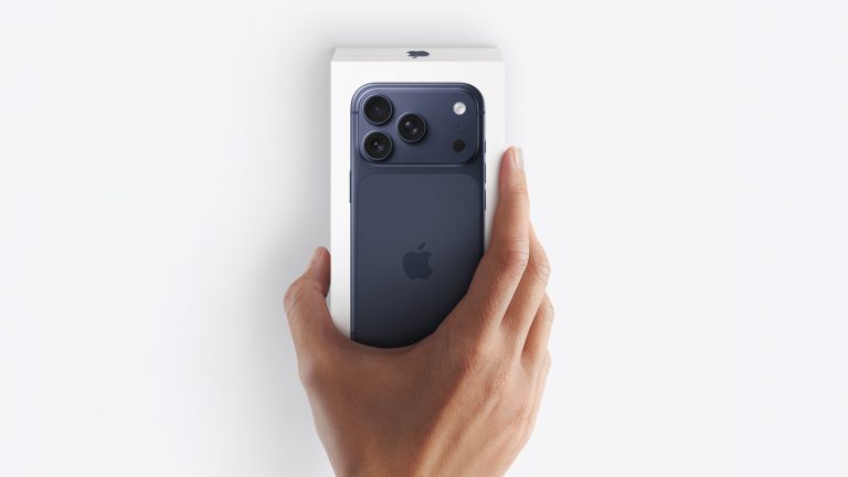 A hand holding a blue iPhone partially out of its white box against a white background, capturing the excitement of an Apple unboxing. The Apple logo and three rear cameras are visible on the phone.