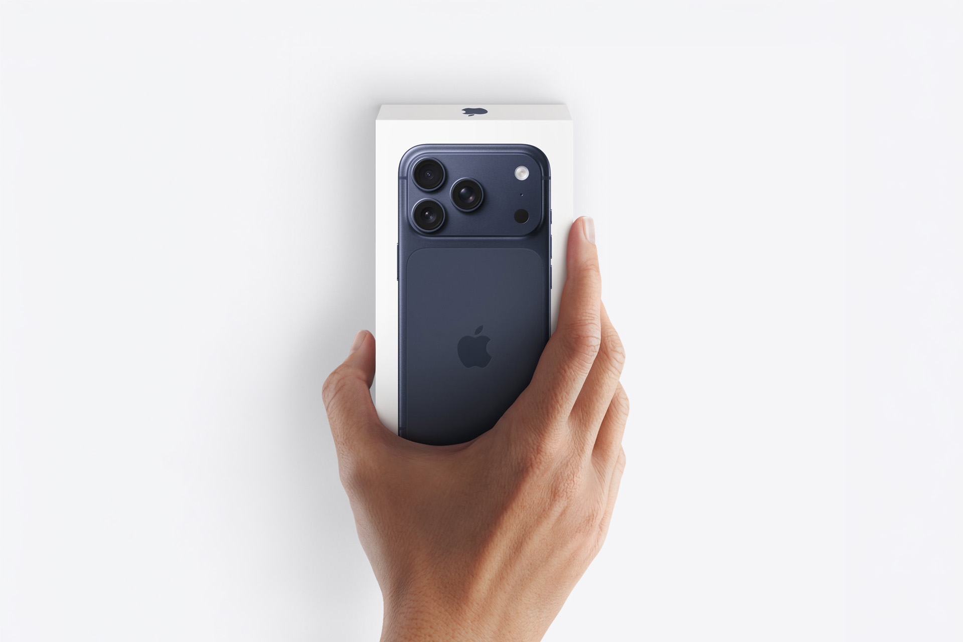 A hand holding a blue iPhone partially out of its white box against a white background, capturing the excitement of an Apple unboxing. The Apple logo and three rear cameras are visible on the phone.
