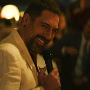 A man in a cream-colored suit smiles while holding a microphone, appearing to speak or perform for an audience in a warmly lit, indoor setting reminiscent of a classic scene from Cape Fear.