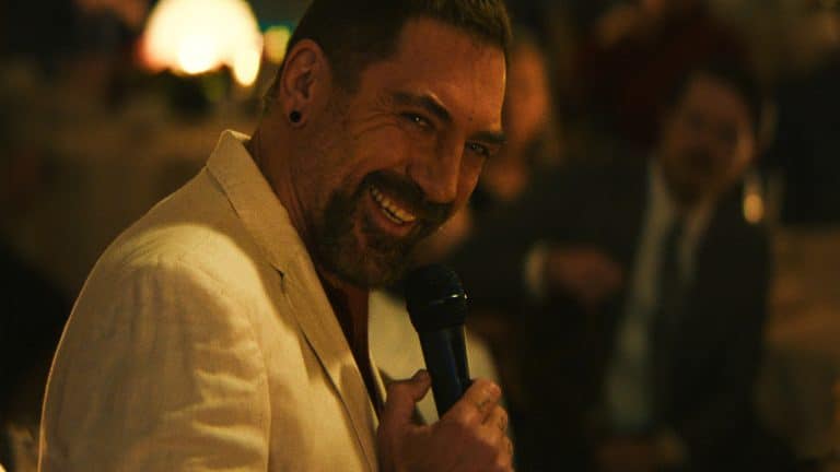 A man in a cream-colored suit smiles while holding a microphone, appearing to speak or perform for an audience in a warmly lit, indoor setting reminiscent of a classic scene from Cape Fear.