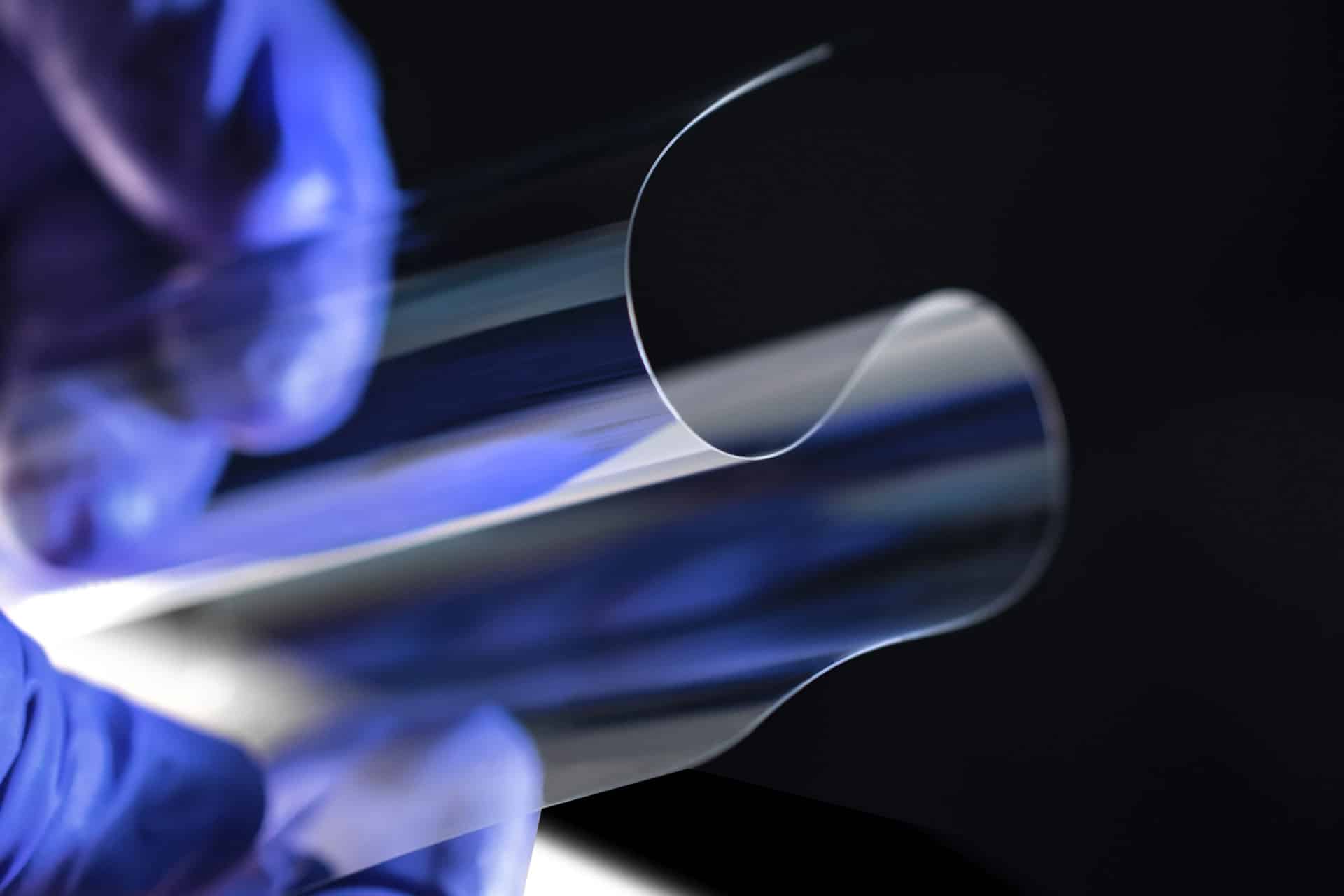 A gloved hand holds a thin, flexible sheet of transparent Apple Corning glass, which bends easily against a dark background.