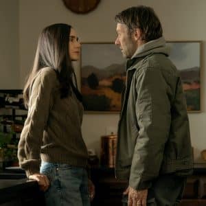 A woman and a man stand facing each other in a kitchen, engaged in a serious conversation. The woman leans against the counter while the man stands close to her, as a Dark Matter Series poster hangs on the wall behind them.