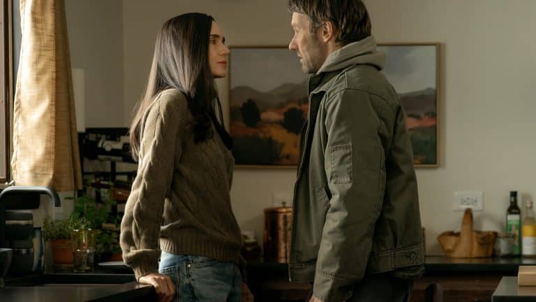 A woman and a man stand facing each other in a kitchen, engaged in a serious conversation. The woman leans against the counter while the man stands close to her, as a Dark Matter Series poster hangs on the wall behind them.