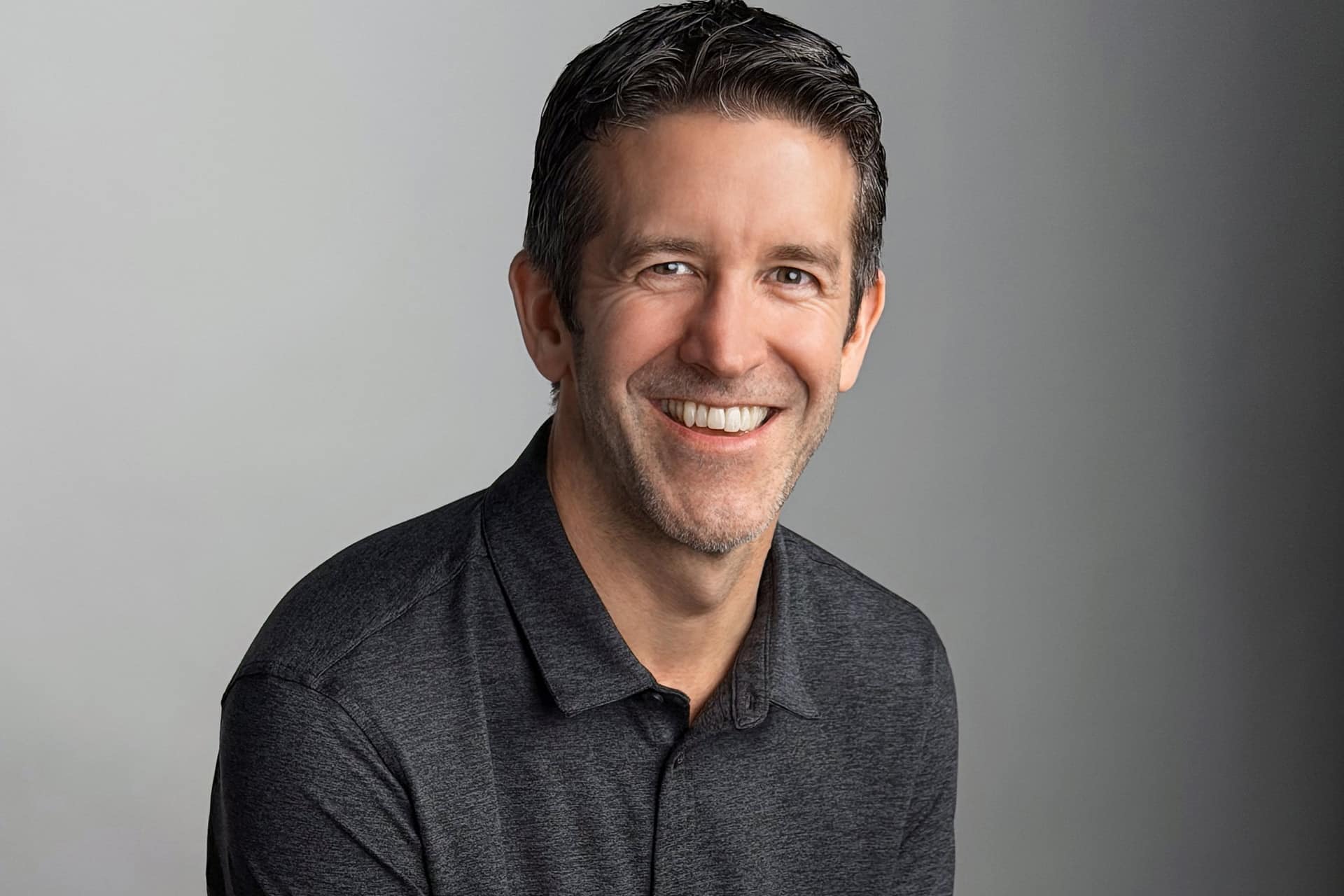 John Ternus - A man with short dark hair and light stubble is smiling at the camera. He is wearing a dark gray collared shirt and is posed against a plain light gray background.