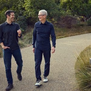 Two men walk side by side on a gravel path surrounded by greenery, smiling and engaged in conversation. Both are casually dressed in long-sleeve shirts, jeans, and sneakers.