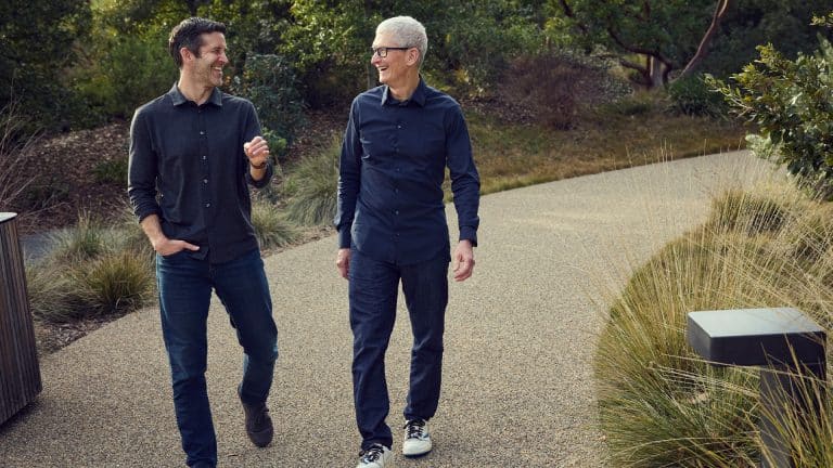 Two men walk side by side on a gravel path surrounded by greenery, smiling and engaged in conversation. Both are casually dressed in long-sleeve shirts, jeans, and sneakers.